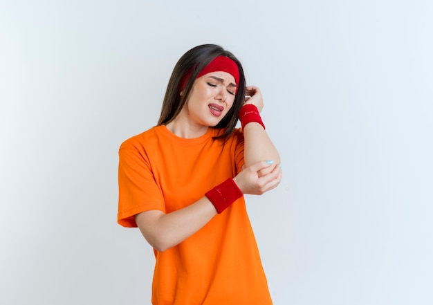 Aching young sporty woman wearing headband and wristbands holding and looking at elbow isolated