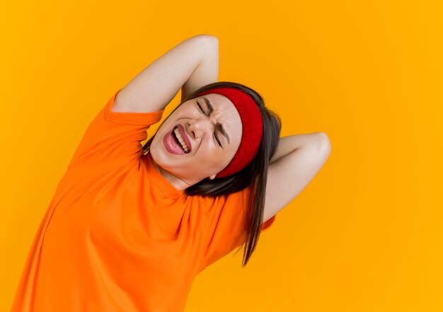 Aching young sporty woman wearing headband and wristbands keeping hands behind back exercising with closed eyes 