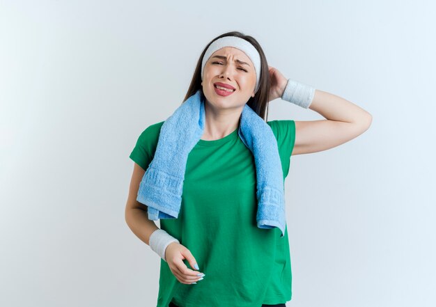 Aching young sporty woman wearing headband and wristbands with towel around neck looking keeping hand behind head 