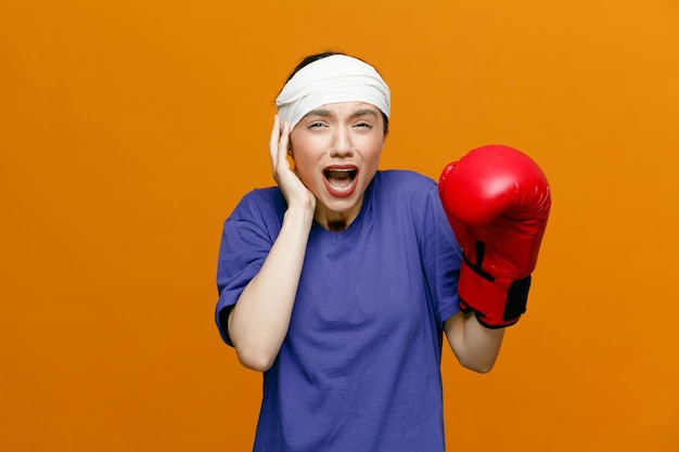 Aching young sporty woman wearing tshirt and boxing glove keeping hand on head looking at camera keeping another hand in air with head wrapped with bandage isolated on orange background