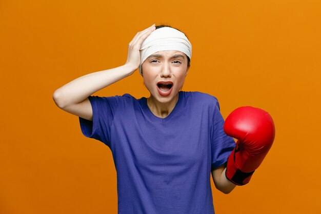 Aching young sporty woman wearing tshirt and boxing glove looking at camera keeping hand on head with head wrapped with bandage isolated on orange background