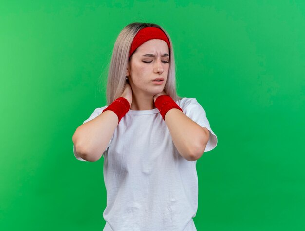 Aching young sporty woman with braces wearing headband and wristbands holds neck with two hands isolated on green wall