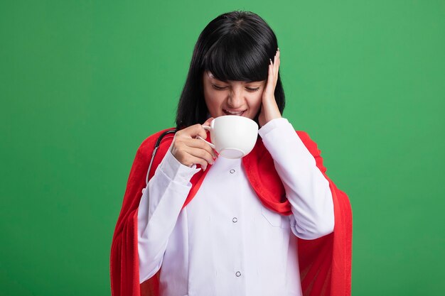 Aching young superhero girl wearing stethoscope with medical robe and cloak drinks tea putting hand on head isolated on green wall