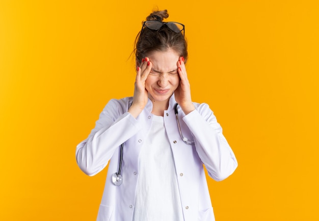 Free photo aching young woman in doctor uniform with stethoscope holding her head