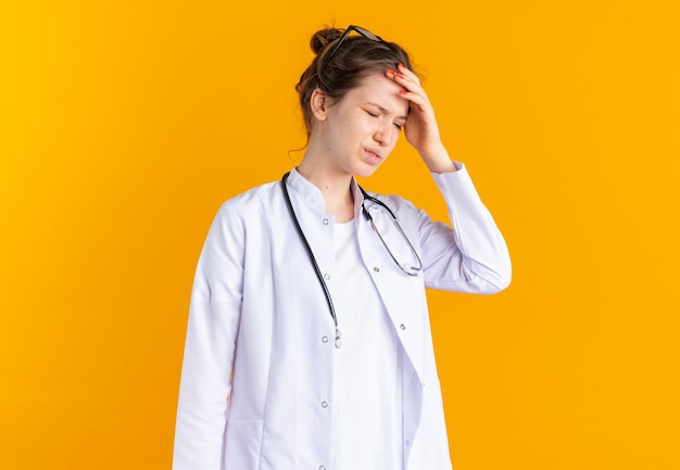 Aching young woman in doctor uniform with stethoscope putting hand on her forehead