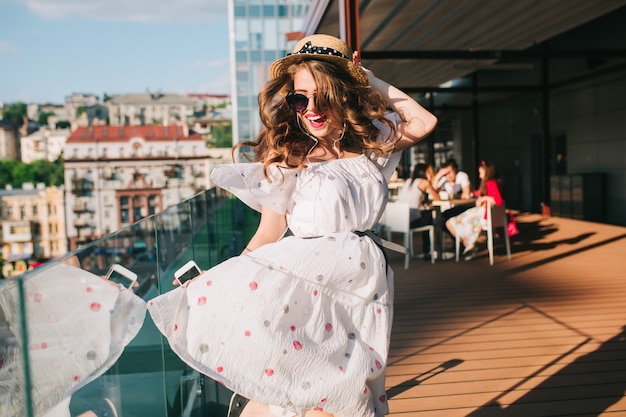 Activ girl  in sunglasses is listening to music through headphones on the terrace. She wears a white dress with bare shoulders, red lipstick and hat . She is dancing like crazy.