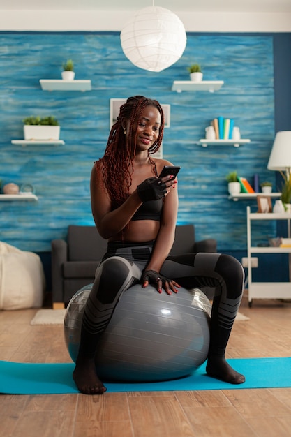 강한 몸과 건강한 라이프 스타일을 얻기 위해 요가 매트에서 운동 한 후 집 거실에서 스위스 공에 앉아 스마트 폰으로 채팅하는 활동적인 운동 여성