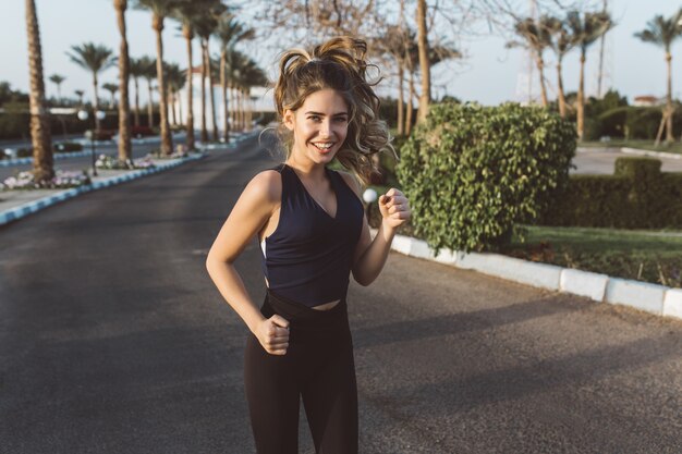 Active attractive slim young woman in sportswear running, smiling on street with palm trees in tropical city. Fashionable model, workout, training, cheerful mood, motivation