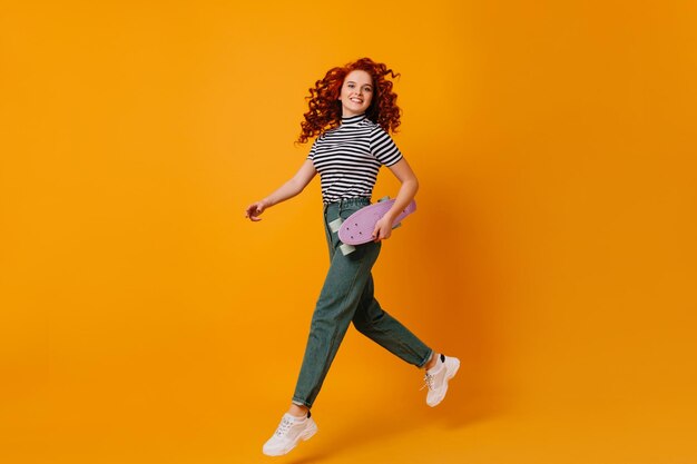Active blueeyed redhead girl in white stylish sneakers and jeans jumping on orange background holding lilac longboard