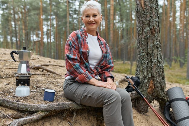 Active cheerful middle aged woman sitting under tree with camping gear boiling water for tea on gas stove burner, having small break during long distance trek. People, adventure, travel and hiking