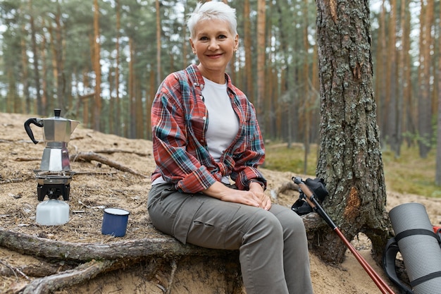 Active cheerful middle aged woman sitting under tree with camping gear boiling water for tea on gas stove burner, having small break during long distance trek. People, adventure, travel and hiking