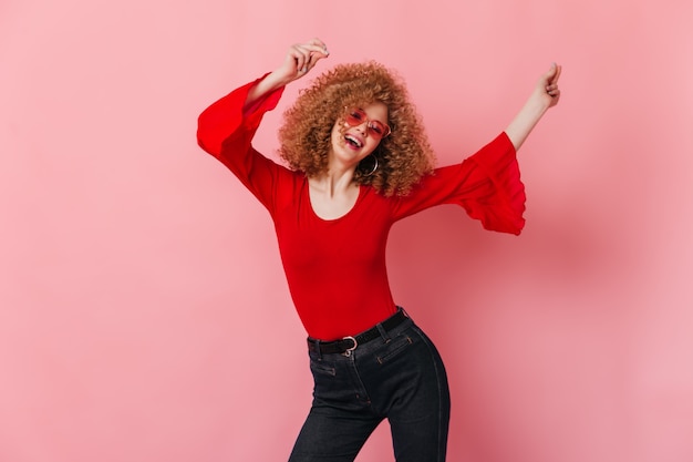 Бесплатное фото Активная кудрявая дама в солнечных очках, красном топе с длинными рукавами и джинсовых брюках смеется и танцует на розовом пространстве.