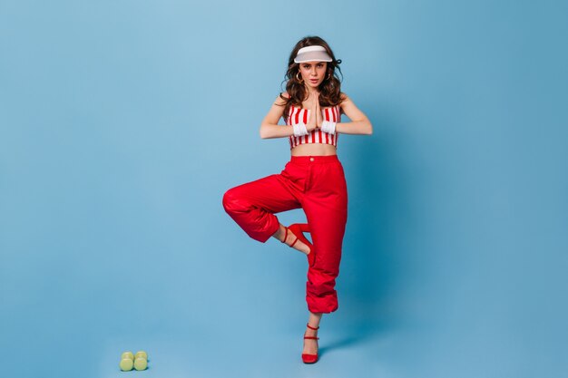 Active curly woman in white cap and red outfit standing in tree pose