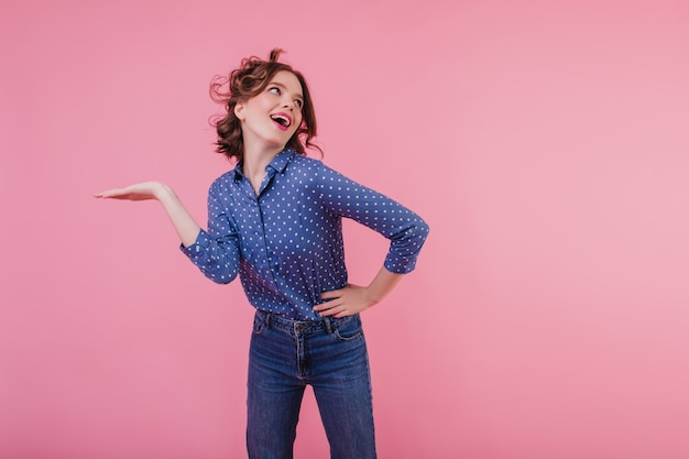 Attiva ragazza dai capelli scuri in camicetta blu che balla foto al coperto di allegra giovane donna con i capelli ondulati che si diverte sul muro rosa.