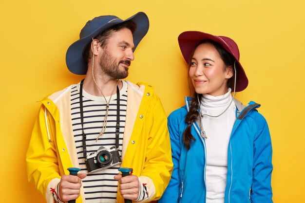 Active diverse woman and man look gladfully at each other, wear raincoat and jacket, hats, explore new place