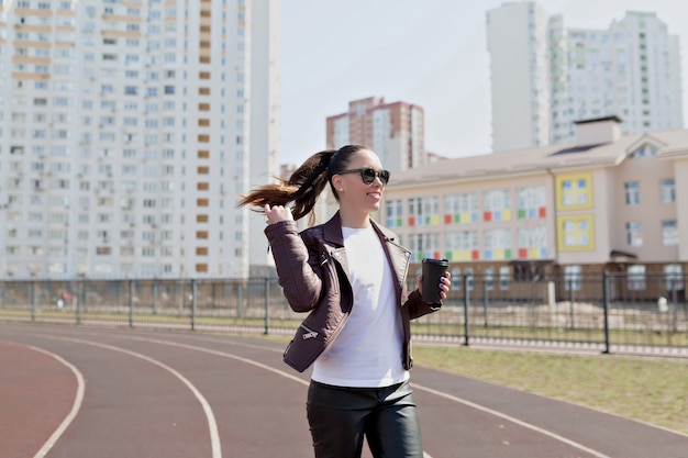 Active excited modern lady dressed leather jacket and white t-shirt and black glasses walks down with coffee cup