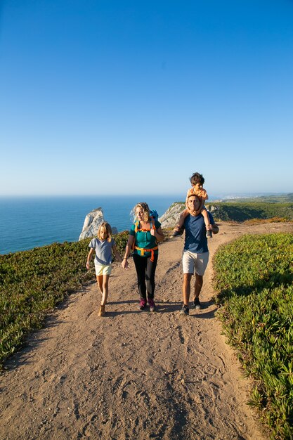 Active family couple and children  hiking along seaside, walking on path. Boy riding on dads neck. Full length. Nature and recreation concept