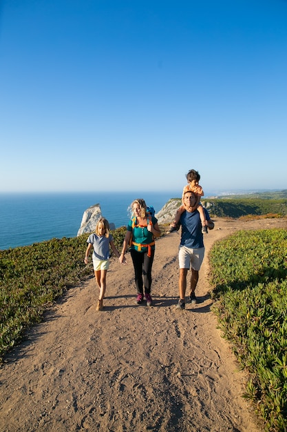 アクティブな家族のカップルや子供たちが海辺に沿ってハイキングし、小道を歩いています。お父さんの首に乗っている少年。全長。自然とレクリエーションの概念