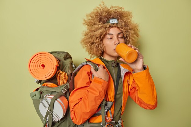 Active female camper feels thirsty drinks water after strolling in mountains carries heavy rucksack dressed in orange windbreaker isolated over green background People and recreation concept