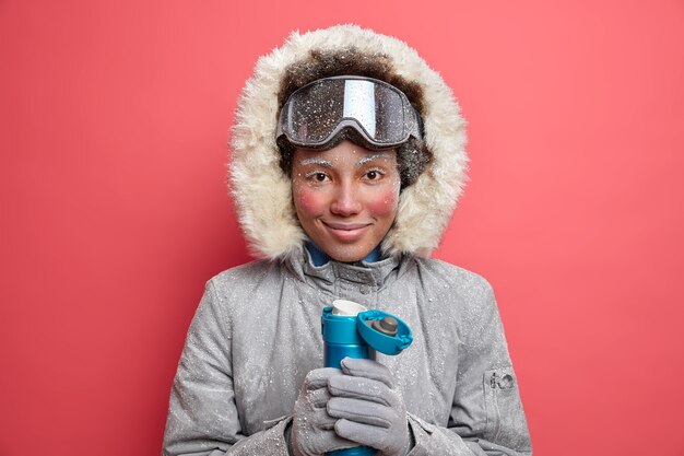 Активная сноубордистка в теплой верхней одежде с красной кожей и замерзшим лицом в холодную зимнюю погоду пьет горячий напиток из термоса.