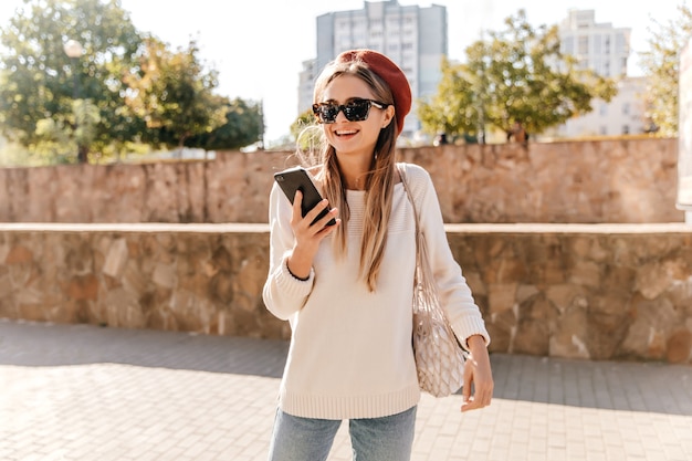 Ragazza francese attiva in occhiali da sole e berretto agghiacciante all'aperto. piacevole donna dai capelli lunghi con il telefono in giro per la città.