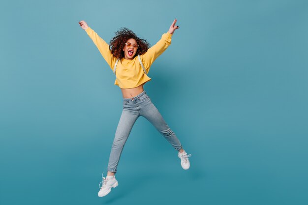 적극적인 소녀는 혀를 보여줍니다. 노란색 스웨터와 청바지에 고립 된 푸른 공간에 행복 점프 여자.