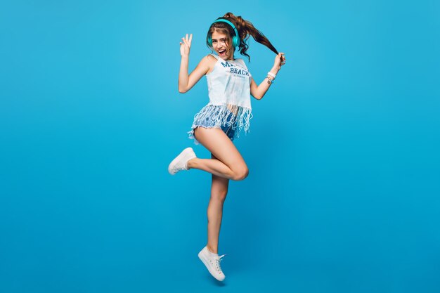 Active girl with long curly hair in tail in jump on blue background in studio. She wears white T-shirt, shorts. She is listening to music with blue headphones.