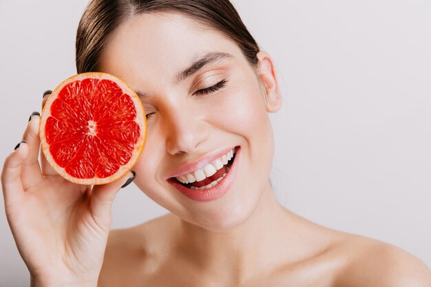 Active girl without makeup posing with grapefruit, bringing energy. Portrait of brunette with snow-white smile on isolated wall.