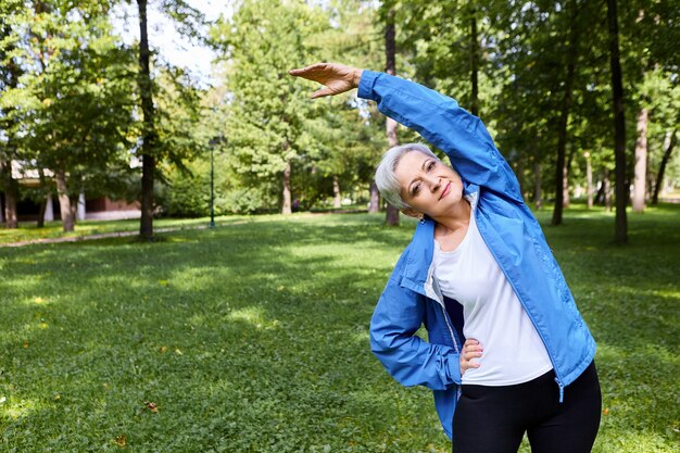 アクティブな白髪のシニア白人女性が片手を腰に持ち、公園でサイドベンドをしながら腕を上げ、有酸素運動の前に体を温め、幸せな楽しい表情をしています