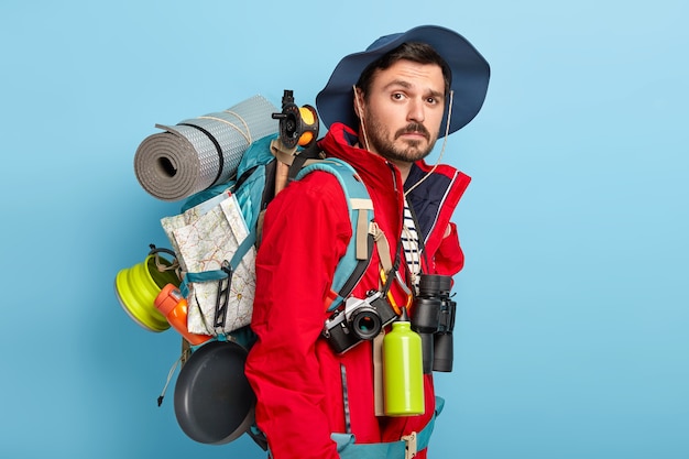 Active handsome man with mustache and bristle, carries tourist rucksack on back, walks in forest, has hiking trip, wears red jacket and hat