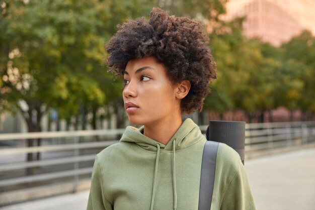 Active healthy woman with curly hair dressed in hoodie carries rolled fitness mat concentrated away thoughtfully strolls outdoors during sunny day returns home after training Sport concept