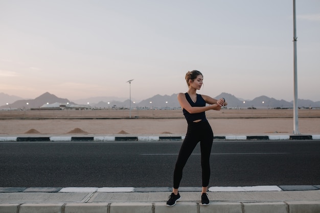 Free photo active joyful sportswoman walking on road on sunset. tropical country, looking at watch on hand, cheerful mood, motivation, workout, healthy lifestyle..