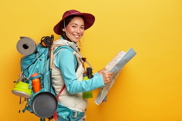 Active korean female tourist carries big rucksack, wears hat and casual clothes, holds map, studies route, has much things needed during traveling
