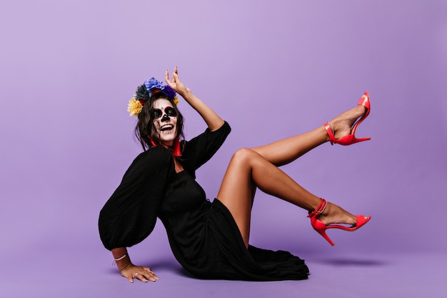 Active lady with crown of blue flowers shows stylish coral-colored high heels. portrait of playful girl with skull mask sitting on floor.