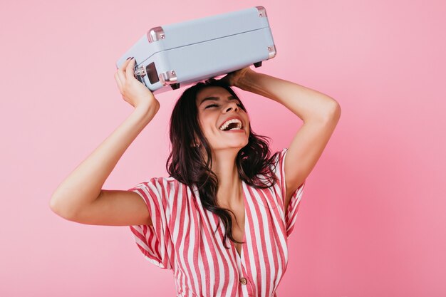 Active lady with dark curls sincerely laughs. Girl in T-shirt with V-neck raised her bag over her head.