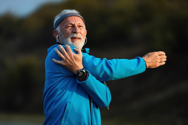 Active mature athlete warming up and stretching his arm while having sports training in nature