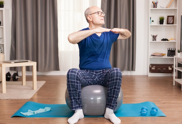 Активный старик занимается спортом в своей уютной квартире, используя швейцарский мяч и коврик для йоги