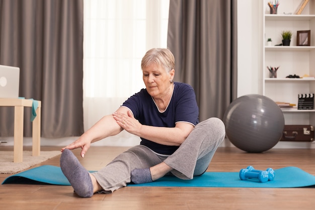 Активная старуха тренируется на коврике для йоги в своей комфортабельной квартире