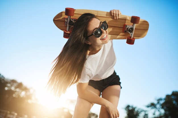 점프할 준비가 된 긴 보드를 들고 카메라에 기대어 있는 여성 스케이트보더의 활동적인 초상화