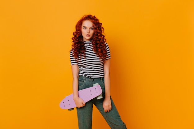 Active redhead girl in stylish denim pants and top posing with skateboard on orange background