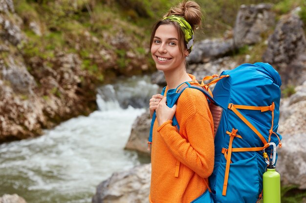 Active rest and positive emotions concept. Cheerful female dressed in casual orange jumper, carries touristic rucksack