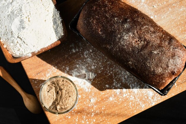 Active rye and wheat sourdough starter in a glass jar next to the ingredient flour and freshly baked whole grain bread Laid out on the table