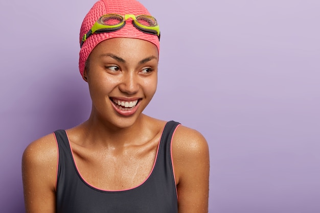 Free photo active smiling dark skinned female swimmer prepares for competition, being wet after diving, dressed in swimming costume, cap and goggles, focused aside, has sporty body. recreation and hobby