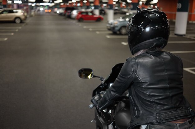 Active way of life, motorcycling, night city and people concept. Rear shot of fashionable confident female biker wearing safety helmet and black leather jacket, riding her motorbike on parking lot
