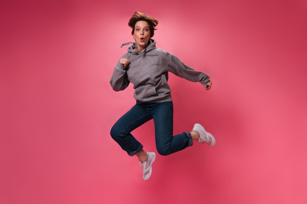 ピンクの背景にジャンプする居心地の良い服装のアクティブな女性。孤立した上を移動するパーカーとジーンズの魅力的な女の子の肖像画