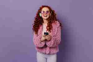 Free photo active woman laughs while listening to music in large headphones. girl in pink woolen jacket and glasses holding phone.