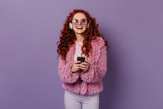 Active woman laughs while listening to music in large headphones. Girl in pink woolen jacket and glasses holding phone.