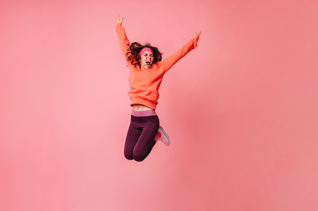 Active woman in orange hoodie and dark leggings vigorously jumping on pink wall