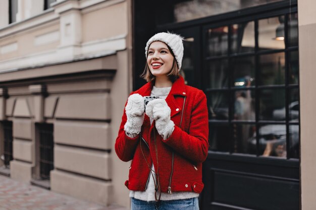 빨간 립스틱과 백설 공주 미소를 가진 활동적인 여성은 복고풍 카메라로 사진을 찍습니다. 밝은 짧은 코트와 장갑을 낀 따뜻한 모자를 쓴 소녀는 산책을 즐깁니다.