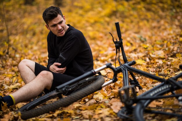 Активный молодой человек, держась за ушибленные или сломанные руки, лежа на тропинке в осеннем лесу на велосипеде